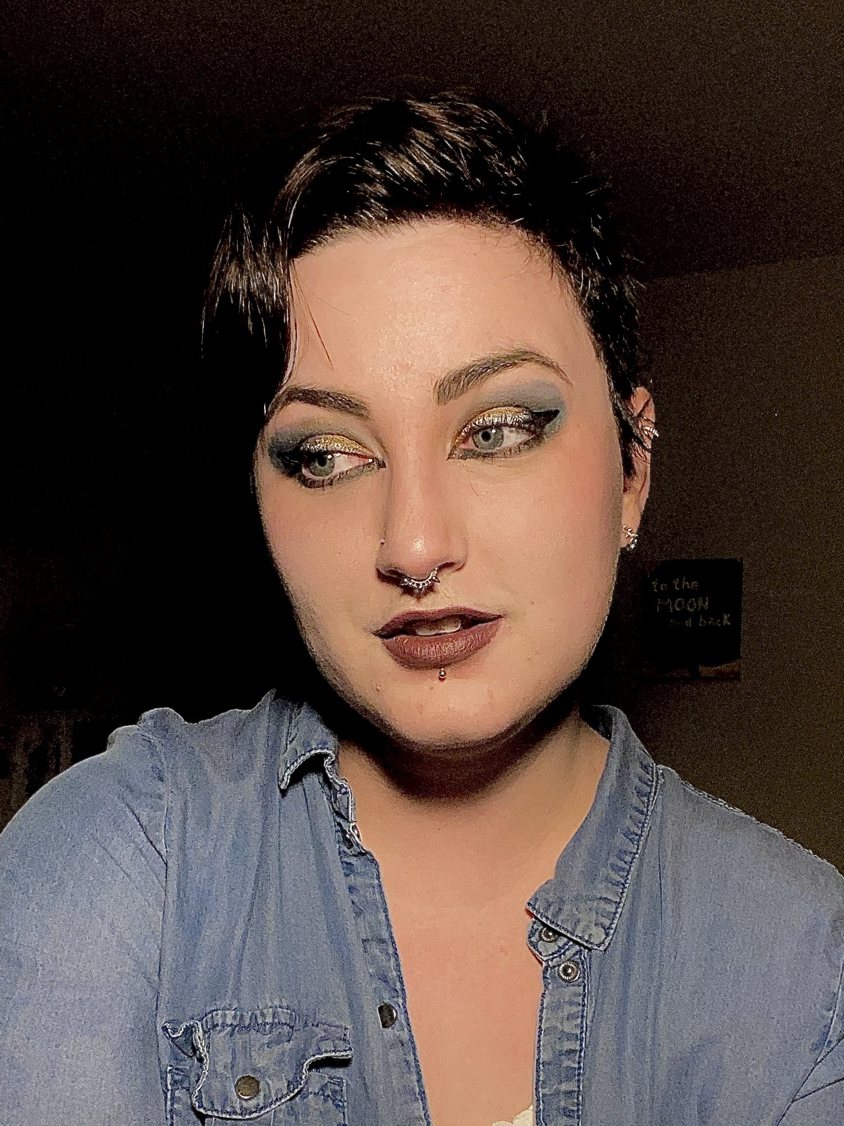 Portrait of Lauren.  Lauren has short brown hair, blue eyes and wears a denim top.  They sport bold eye makeup.  There is bronze shadow on their eyelids surrounded by blue eye shadow spreading out.  Shimmery white lines each upper lid.  Black eyeliner juts out from the outer corners and underlines the eyes, coming to a point below each tear duct.